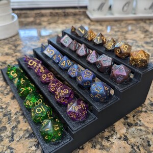 Pode incluir: Um tabuleiro de dados preto de v&aacute;rios n&iacute;veis com uma variedade de dados coloridos e iridescentes com n&uacute;meros dourados. Os dados est&atilde;o dispostos em filas no tabuleiro, com os n&uacute;meros mais altos na parte superior e os n&uacute;meros mais baixos na parte inferior.