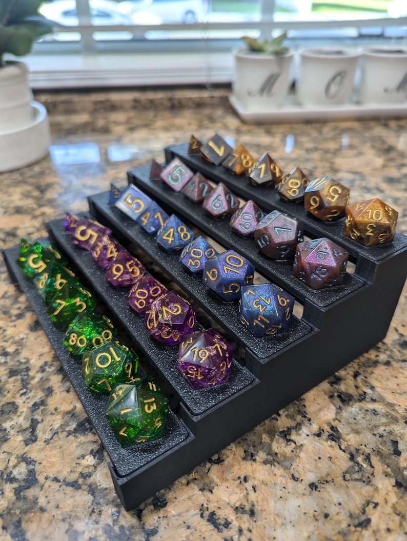 Pode incluir: Um tabuleiro de dados preto de v&aacute;rios n&iacute;veis com uma variedade de dados coloridos e iridescentes com n&uacute;meros dourados. Os dados est&atilde;o dispostos em filas no tabuleiro, com os n&uacute;meros mais altos na parte superior e os n&uacute;meros mais baixos na parte inferior.