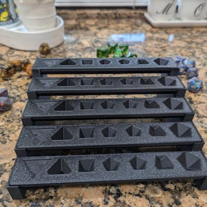 Pode incluir: Uma bandeja de dados de pl&aacute;stico preto com cinco n&iacute;veis, cada um com m&uacute;ltiplos compartimentos para guardar dados. A bandeja &eacute; projetada para conter uma variedade de tamanhos e formas de dados.