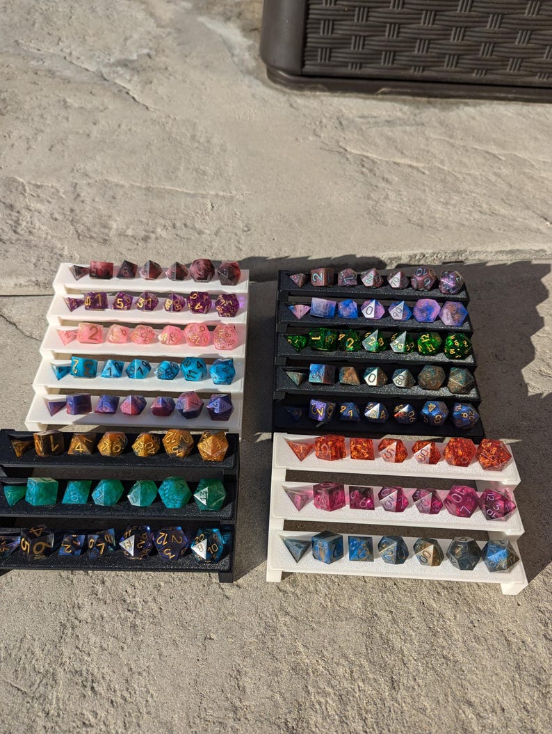 Pode incluir: Um conjunto de quatro suportes de exibi&ccedil;&atilde;o em camadas de acr&iacute;lico branco com filas de dados coloridos. Os dados s&atilde;o feitos de resina e t&ecirc;m uma variedade de cores e padr&otilde;es, incluindo rosa, azul, verde e dourado. Os dados s&atilde;o dispostos em filas nos suportes, com cada fila contendo uma cor ou padr&atilde;o diferente.