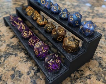 May include: A black plastic tiered display stand with three levels, holding a collection of colorful dice with gold and silver accents. The dice are arranged in rows on each level, with the numbers facing up.