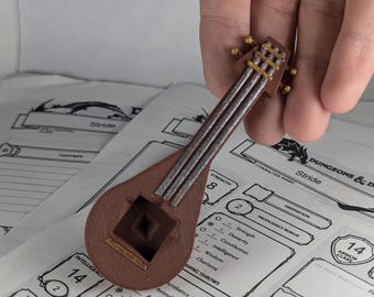 Pode incluir: Um alaúde em miniatura de madeira marrom com detalhes dourados e cordas prateadas. O alaúde está sobre uma superfície branca com uma folha de personagem de Dungeons and Dragons no fundo.