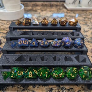 Pode incluir: Um conjunto de 10 dados poli&eacute;dricos em uma caixa de armazenamento de pl&aacute;stico preto. Os dados s&atilde;o feitos de resina transparente verde e azul com glitter dourado e n&uacute;meros dourados. Os dados s&atilde;o organizados em filas na caixa.
