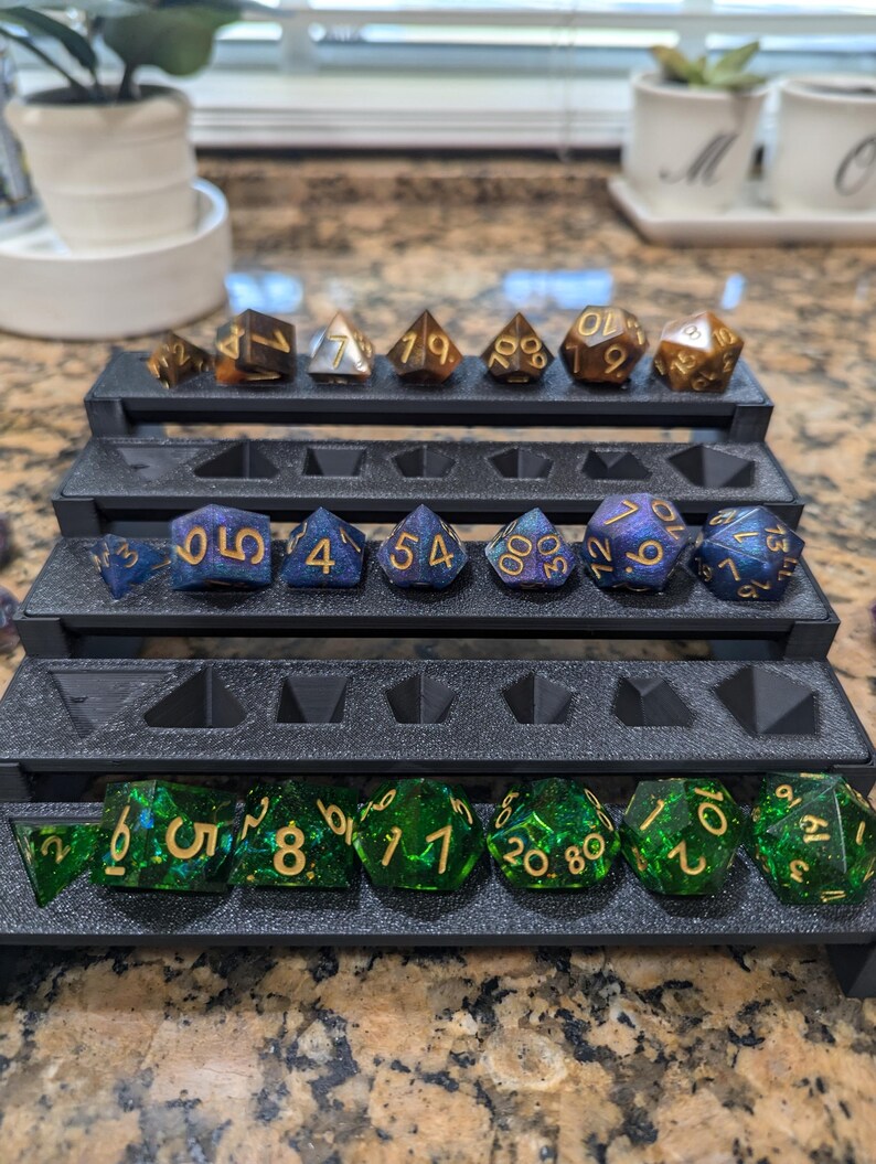 Pode incluir: Um conjunto de 10 dados poli&eacute;dricos em uma caixa de armazenamento de pl&aacute;stico preto. Os dados s&atilde;o feitos de resina transparente verde e azul com glitter dourado e n&uacute;meros dourados. Os dados s&atilde;o organizados em filas na caixa.