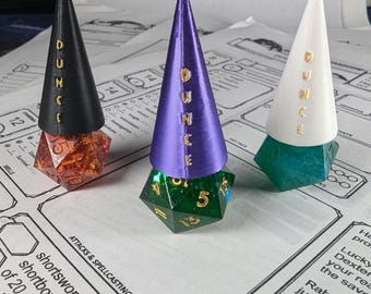 Pode incluir: Três torres de dados em forma de cone impressas em 3D em preto, roxo e branco com a palavra "DUNCE" impressa em cada uma. Cada torre tem um dado d6 verde, vermelho e azul no topo.