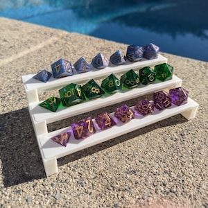 May include: A white three-tiered display stand with a variety of iridescent dice in shades of blue, green, and purple. The dice have gold numbers and are arranged in rows on the stand.