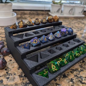 Pode incluir: Uma torre de dados de pl&aacute;stico preto com cinco n&iacute;veis. Cada n&iacute;vel tem ranhuras para dados. A torre est&aacute; cheia de dados de v&aacute;rias cores com n&uacute;meros dourados. Os dados s&atilde;o uma mistura de verde, azul e marrom.