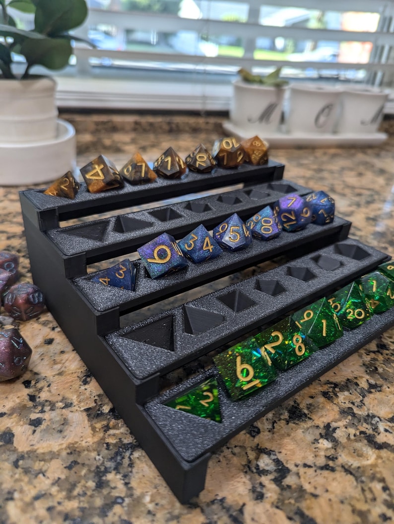 Pode incluir: Uma torre de dados de pl&aacute;stico preto com cinco n&iacute;veis. Cada n&iacute;vel tem ranhuras para dados. A torre est&aacute; cheia de dados de v&aacute;rias cores com n&uacute;meros dourados. Os dados s&atilde;o uma mistura de verde, azul e marrom.
