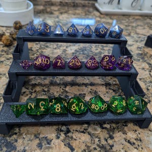 Pode incluir: Um conjunto de 18 dados em um suporte de exibi&ccedil;&atilde;o de tr&ecirc;s n&iacute;veis. Os dados s&atilde;o feitos de resina transparente com glitter dourado e n&uacute;meros dourados. Os dados s&atilde;o dispostos em tr&ecirc;s filas, com seis dados em cada fila. Os dados s&atilde;o de v&aacute;rias formas e tamanhos, incluindo d4, d6, d8, d10, d12 e d20.