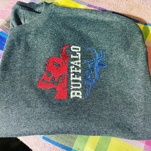 Puede incluir: Una camiseta gris oscura con un diseño bordado de una cabeza de búfalo roja y una silueta azul de un ciervo con la palabra "BUFFALO" en letras blancas.