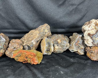May include: A collection of raw, unpolished agate rocks in various shapes and sizes. The stones feature earthy tones of brown, tan, and grey, with some pieces displaying vibrant orange, red, and white crystalline banding and translucent quartz centers.