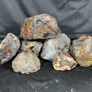 Op de afbeelding: Een verzameling van elf ruwe, ongepolijste rotsen en mineralen in verschillende vormen en maten. De stenen hebben aardse tinten zoals bruin, beige, grijs en roestoranje, waarbij sommige stenen doorschijnende, kristallijne en getextureerde oppervlakken vertonen.