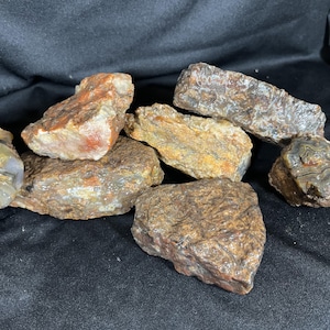May include: A collection of raw, unpolished rocks featuring varied textures and earthy tones. The stones display shades of brown, tan, orange, and grey, with some showing crystalline surfaces, layered patterns, and rough, jagged edges throughout.