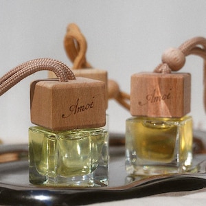 May include: Two small, square glass bottles filled with yellow liquid, topped with wooden caps and tan cords. The wooden caps are engraved with the word "Amoi". The bottles are displayed on a silver tray, suggesting a fragrance or essential oil product.