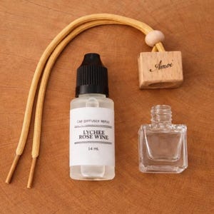 May include: A car air freshener set with a clear glass bottle, a refill bottle labeled "Lychee Rose Wine", a small wooden block with "Amoi" printed on it, and a yellow cord. The set is on a wooden surface.