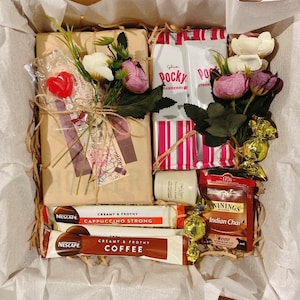 May include: A gift basket filled with various items, including a book wrapped in brown paper with a red heart, a box of Pocky strawberry snacks, a box of Twinings Indian Chai tea, a jar of hand cream, and two Nescafe coffee sachets. The basket is lined with white tissue paper and decorated with pink and white flowers.