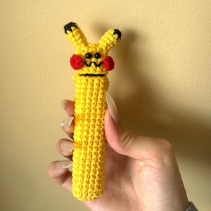 Puede incluir: Una figura de Pikachu amarilla hecha a mano con ganchillo, con ojos negros, boca en zigzag negra, mejillas rojas y orejas con puntas negras. La figura es cil&iacute;ndrica y se sostiene en una mano.