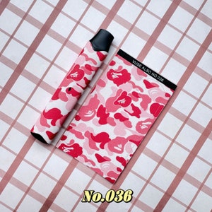以下が含まれることがあります： ピンクと赤のカモフラージュ柄のVAPEペンとマッチするケース。ケースには「VUSE ALSO NO. 036」の文字がプリントされています。VAPEペンには黒いマウスピースが付いています。背景はピンクと白のチェック柄です。
