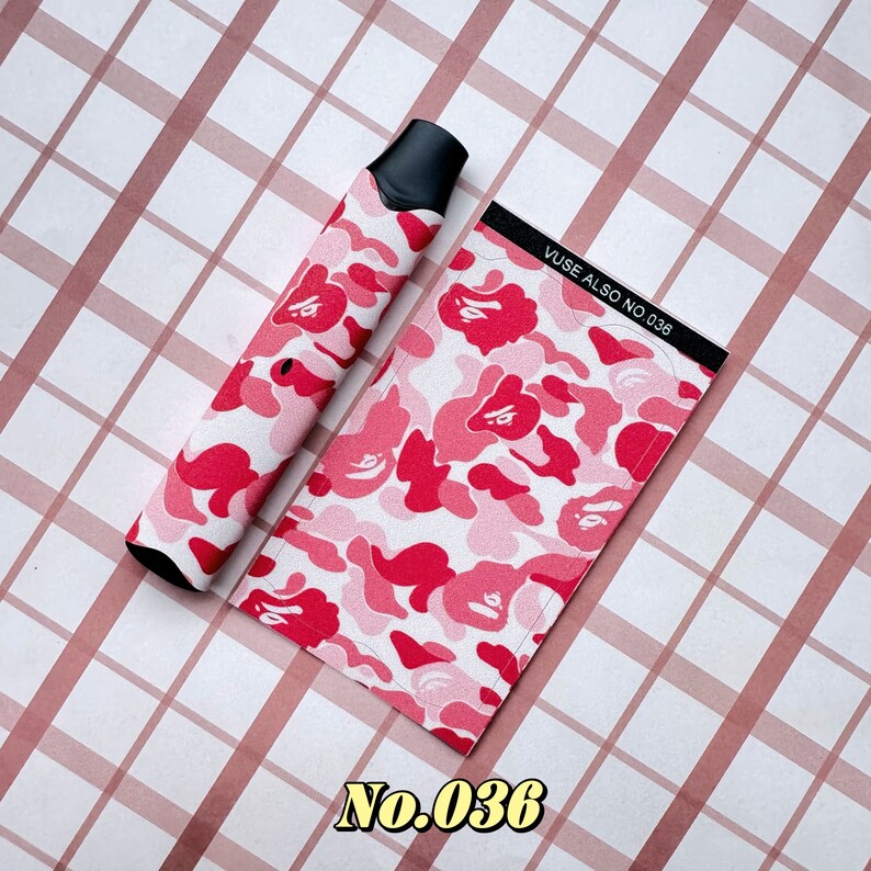 以下が含まれることがあります： ピンクと赤のカモフラージュ柄のVAPEペンとマッチするケース。ケースには「VUSE ALSO NO. 036」の文字がプリントされています。VAPEペンには黒いマウスピースが付いています。背景はピンクと白のチェック柄です。