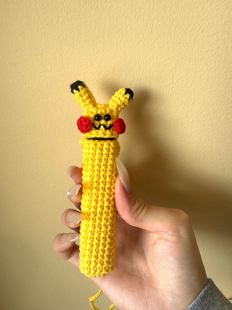 Puede incluir: Una figura de Pikachu amarilla hecha a mano con ganchillo, con ojos negros, boca en zigzag negra, mejillas rojas y orejas con puntas negras. La figura es cil&iacute;ndrica y se sostiene en una mano.