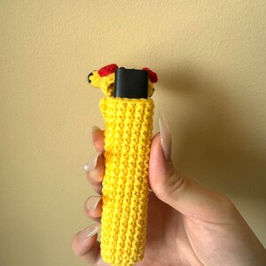 Puede incluir: Una funda de ganchillo amarilla con una peque&ntilde;a cabeza decorativa con detalles rojos. La funda est&aacute; dise&ntilde;ada para contener un objeto rectangular negro. Una cuerda amarilla est&aacute; unida a la parte inferior de la funda.