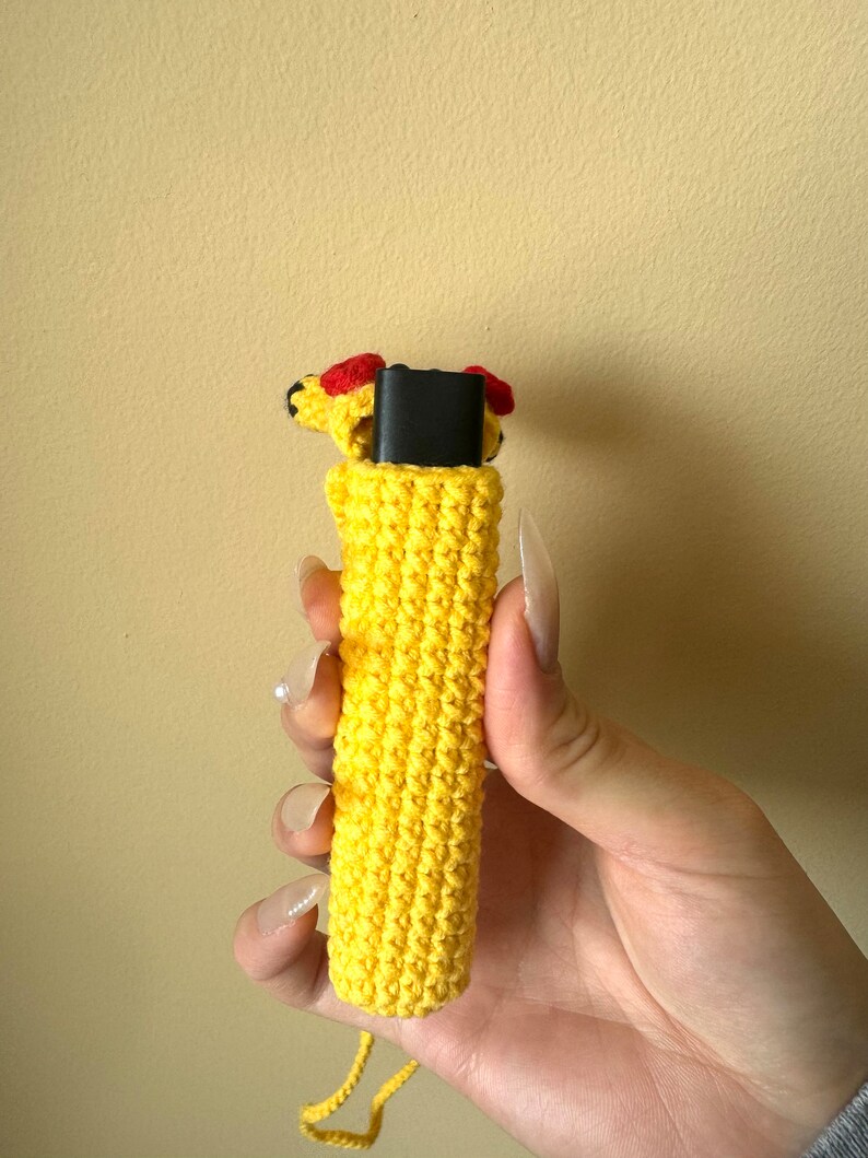 Puede incluir: Una funda de ganchillo amarilla con una peque&ntilde;a cabeza decorativa con detalles rojos. La funda est&aacute; dise&ntilde;ada para contener un objeto rectangular negro. Una cuerda amarilla est&aacute; unida a la parte inferior de la funda.