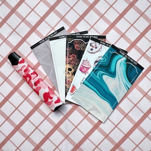 以下が含まれることがあります： 赤とピンクのカモフラージュ柄の電子タバコと、いくつかの装飾的なカードデザイン。カードには、スカル、花柄、抽象的なデザインなど、さまざまなパターンが描かれています。カードには「VUSE ALTO NO.031」から「035」までラベルが貼られています。