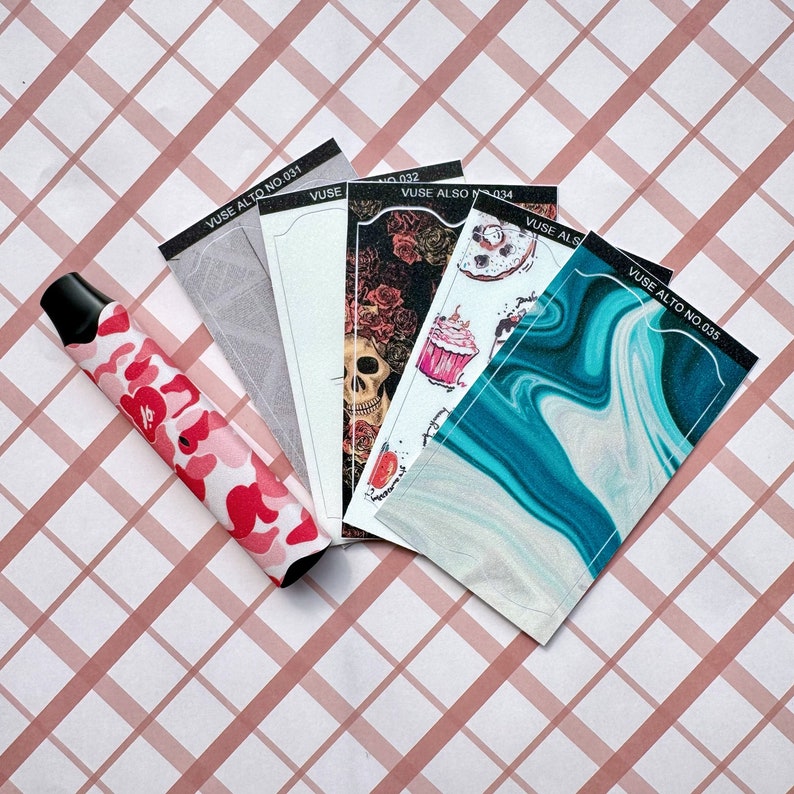 以下が含まれることがあります： 赤とピンクのカモフラージュ柄の電子タバコと、いくつかの装飾的なカードデザイン。カードには、スカル、花柄、抽象的なデザインなど、さまざまなパターンが描かれています。カードには「VUSE ALTO NO.031」から「035」までラベルが貼られています。