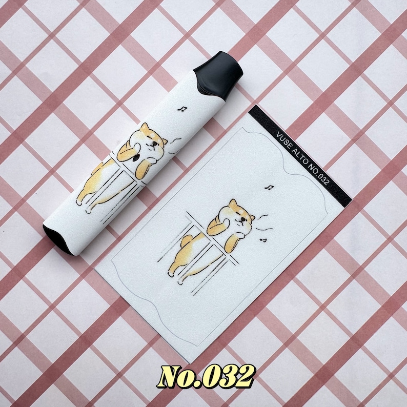 以下が含まれることがあります： 黒い先端が付いた白いVAPEペンと、それに合わせたカード。どちらも、音符が付いた漫画の柴犬のデザインが特徴です。カードには「VUSE ALTO NO.032」と書かれています。背景はピンクと白のチェック柄です。