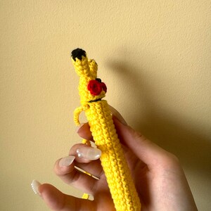 Puede incluir: Un bol&iacute;grafo de ganchillo amarillo hecho a mano inspirado en Pikachu. El bol&iacute;grafo presenta orejas con puntas negras, mejillas rojas y una nariz negra. El bol&iacute;grafo se sostiene en una mano sobre un fondo amarillo. Es un art&iacute;culo &uacute;nico y artesanal.