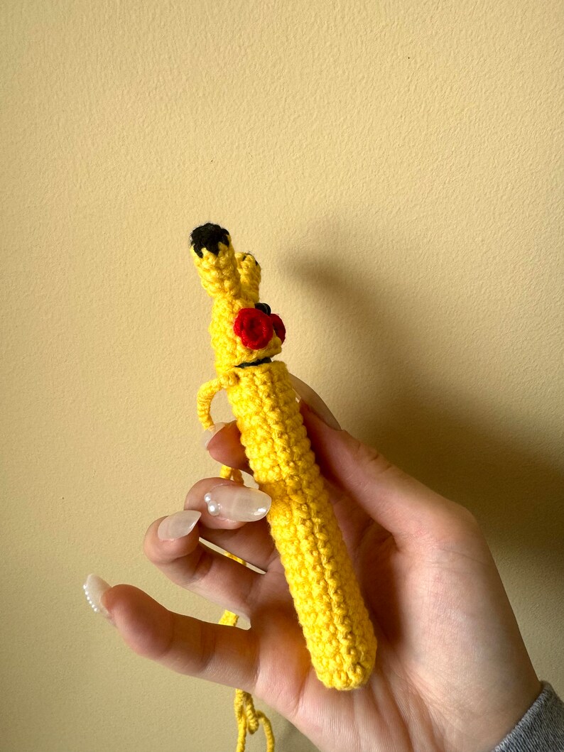 Puede incluir: Un bol&iacute;grafo de ganchillo amarillo hecho a mano inspirado en Pikachu. El bol&iacute;grafo presenta orejas con puntas negras, mejillas rojas y una nariz negra. El bol&iacute;grafo se sostiene en una mano sobre un fondo amarillo. Es un art&iacute;culo &uacute;nico y artesanal.
