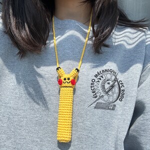 Puede incluir: Una funda de tel&eacute;fono amarilla de ganchillo con un dise&ntilde;o de Pikachu, mejillas rojas y detalles negros, sujeta a un cord&oacute;n amarillo. La persona lleva una sudadera gris con el texto "ELECTRO M&Eacute;CANIQUE CFC 2000".