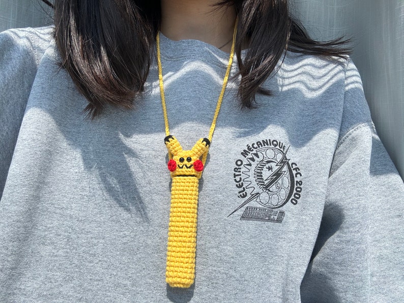 Puede incluir: Una funda de tel&eacute;fono amarilla de ganchillo con un dise&ntilde;o de Pikachu, mejillas rojas y detalles negros, sujeta a un cord&oacute;n amarillo. La persona lleva una sudadera gris con el texto "ELECTRO M&Eacute;CANIQUE CFC 2000".