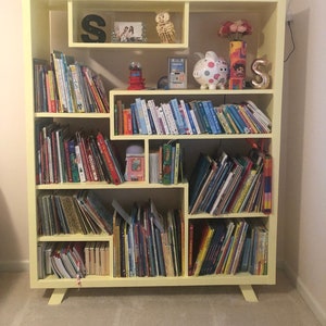 Puede incluir: Una estantería de color amarillo claro llena de libros y objetos decorativos. La estantería tiene un diseño único con estantes de varios tamaños. Los objetos decorativos incluyen una hucha, una jirafa y fotos enmarcadas.