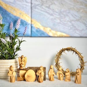 May include: A nativity scene set carved from olive wood. The set includes figures of Mary, Joseph, baby Jesus, an angel, a shepherd, and a donkey. The figures are arranged around a wooden log with a circular piece of wood representing a manger. A wreath of thorns is in the background.