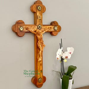 May include: Wooden crucifix with a carved figure of Jesus. The cross is adorned with decorative accents and a sunburst design. The text "The Good Shepherds of Bethlehem" is visible. A white orchid plant in a white pot is in the foreground.
