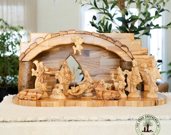 May include: A hand-carved olive wood nativity scene with a detailed archway and figures of Mary, Joseph, baby Jesus, an angel, shepherds, and animals. The scene is set on a wooden base with the words "The Good Shepherds of Bethlehem" inscribed on a circular seal.