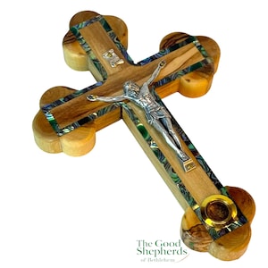 May include: Wooden cross with a silver Jesus figure. The cross features a light brown wood grain, inlaid with iridescent abalone shell and a gold-colored metal accent. The cross has rounded edges.