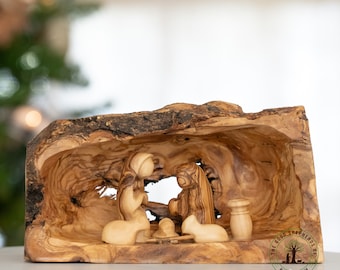 May include: A wooden nativity scene carved from olive wood. The scene depicts Mary, Joseph, and the baby Jesus in a manger. The figures are hand-carved and have a rustic, natural look. The scene is set in a hollowed-out piece of olive wood, which adds to the natural and rustic feel. The scene is surrounded by a circular stamp that reads "The Good Shepherds of Bethlehem".
