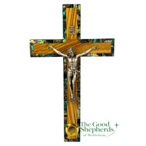 May include: Wooden crucifix with a silver figure of Jesus Christ. The cross features a wooden grain design with iridescent abalone shell accents. The text "INRI" is visible above the figure. The Good Shepherds of Bethlehem logo is in the lower right corner.