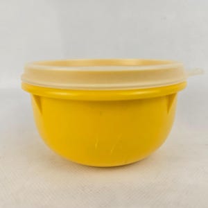 Könnte beinhalten: Eine kleine, gelbe Plastikschüssel mit einem hellbraunen Deckel. Die Schüssel hat eine abgerundete Form und eine glatte Oberfläche. Der Deckel passt fest auf die Oberseite, mit einer kleinen Lasche zum einfachen Entfernen. Die Schüssel ist wahrscheinlich für die Aufbewahrung von Lebensmitteln konzipiert.