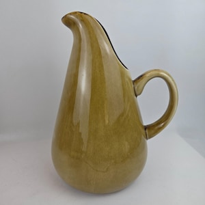 May include: A vintage, teardrop-shaped pitcher with a curved spout and a rounded handle. The pitcher is a muted olive green color with a glossy finish. The interior of the spout is dark brown. The pitcher is likely made of ceramic or stoneware.