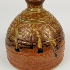 May include: A small, round, brown ceramic vase with a narrow neck. The vase has a speckled glaze with brown, orange, and gold accents. The vase is decorated with a pattern of lines and dots.