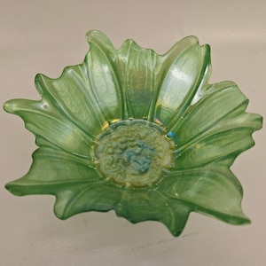 May include: A green glass bowl shaped like a flower. The bowl has a textured surface and a detailed center. The petals are a light green color, and the center has a blue and yellow hue. The bowl is likely used for decorative purposes.