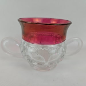 Puede incluir: Una taza de té de cristal transparente con dos asas y un borde rojo rubí. La taza tiene un patrón decorativo grabado alrededor del centro y un ribete dorado en el borde. Las asas son claras y redondeadas.
