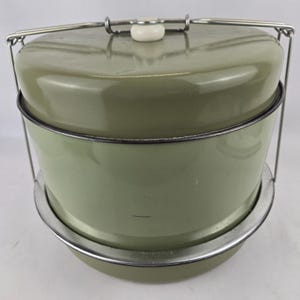 May include: A vintage olive green metal food carrier with a lid and a white handle. The carrier has a silver metal handle and a base. The carrier is designed to keep food warm or cold.