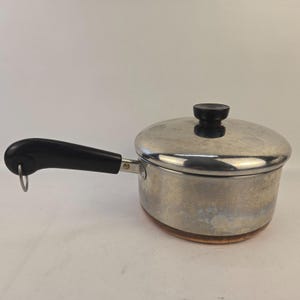 Puede incluir: Una cacerola de acero inoxidable con tapa y asa negra. La olla tiene un fondo color cobre y un pomo negro en la tapa. El asa tiene un lazo metálico para colgar. Diseñada para cocinar.