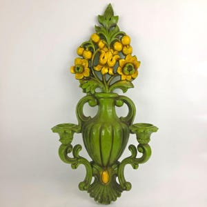 May include: A wall-mounted candle holder featuring a central green vase shape with two side arms for candles. The top section displays a relief carving of yellow flowers and green leaves. The entire piece is finished in a solid green color.