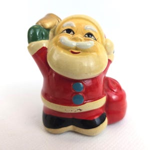 Può includere: Piccola statuina vintage di Babbo Natale. La figura è dipinta con un cappotto rosso, stivali neri e un viso e una barba color crema. Babbo Natale tiene in mano un sacco d'oro e un guanto verde. Due bottoni blu adornano la parte anteriore del cappotto. Una decorazione festiva.