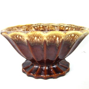 Pode incluir: Uma taça de cerâmica com base de pedestal, apresentando um esmalte castanho escuro na parte inferior e um esmalte de gotejamento em tom creme mais claro ao longo da borda recortada. A taça tem um design exterior canelado e estriado.